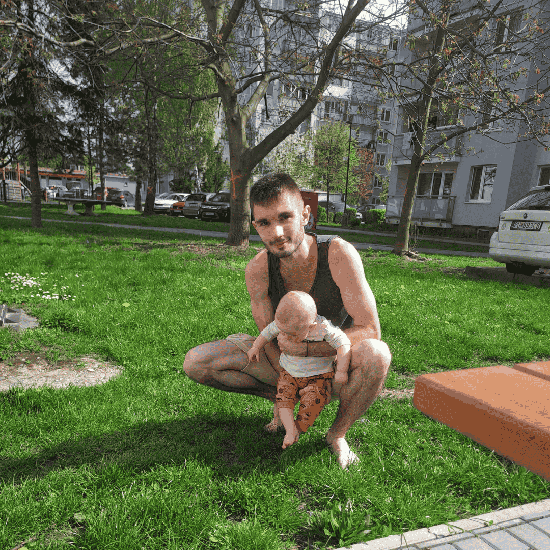 Muž s dieťaťom v tráve, sídlisko v pozadí.